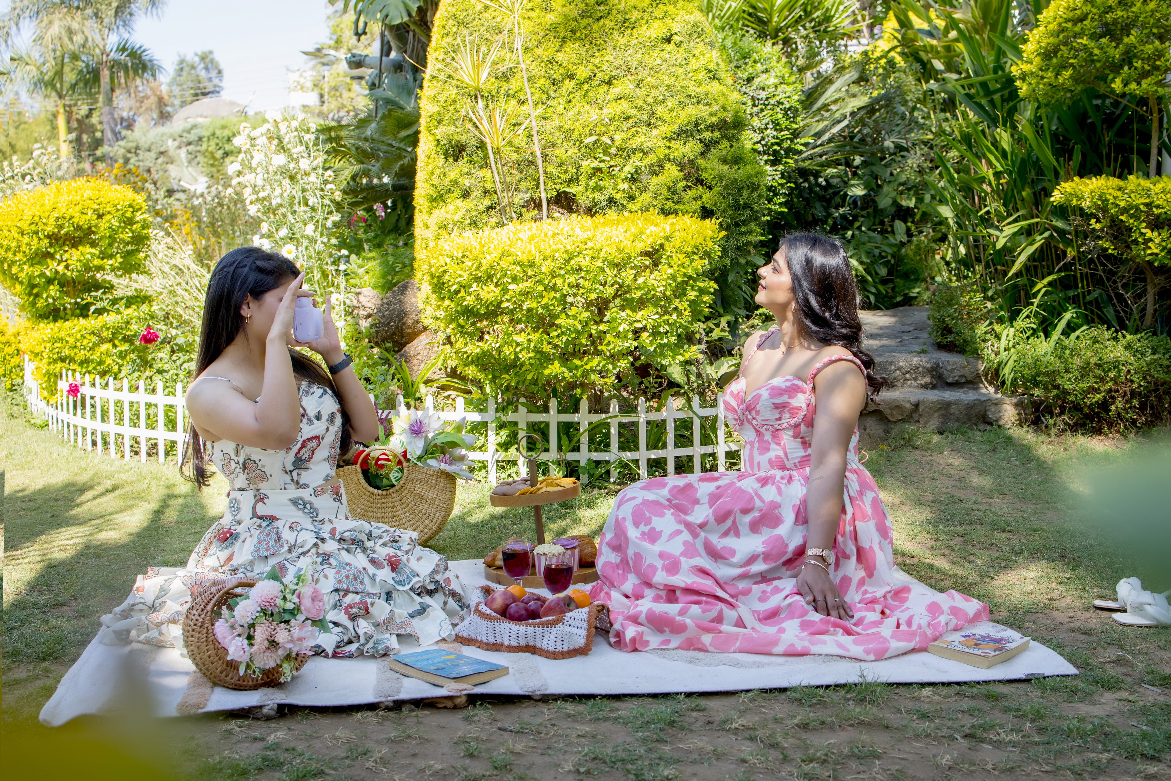Two women in floral dresses sitting on a picnic blanket in a garden.